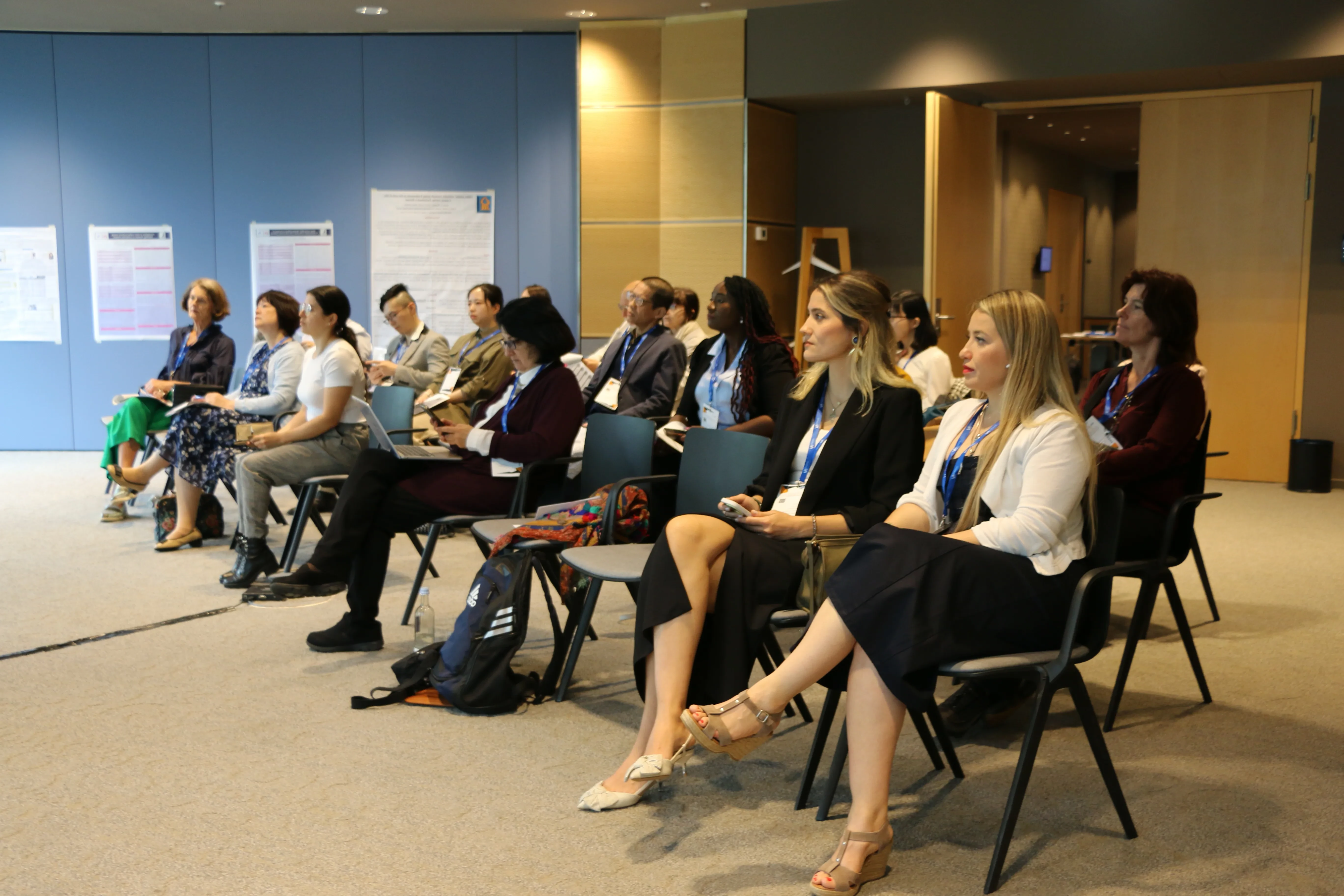 Conference participants with lanyards seated in rows, listening to a session, with research posters on the wall behind them